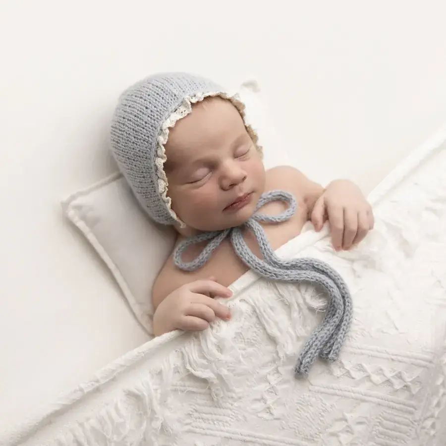 Newborn in a grey knit bonnet asleep on a little pillow under a white knit blanket — newborn photography by Gitty Reiss, Boro Park, Brooklyn