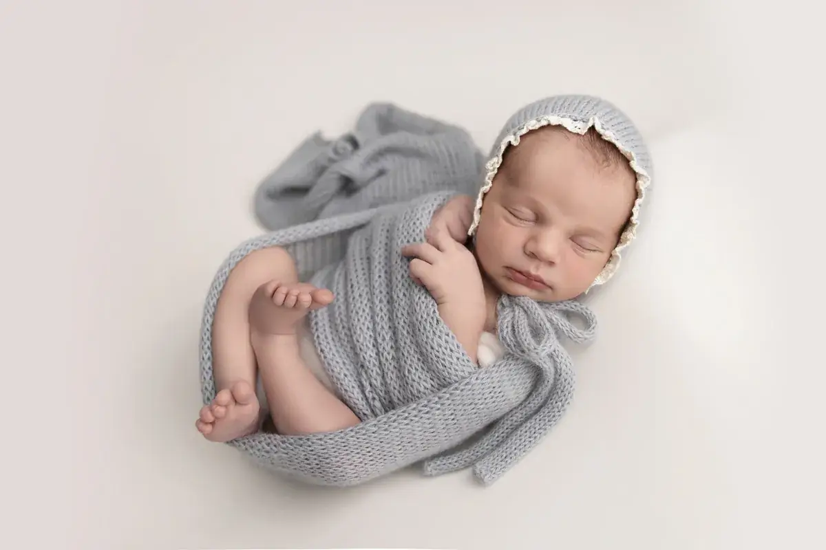Sleeping newborn in a grey knit bonnet and wrap, curled up on a cream backdrop — newborn photography by Gitty Reiss, Boro Park, Brooklyn