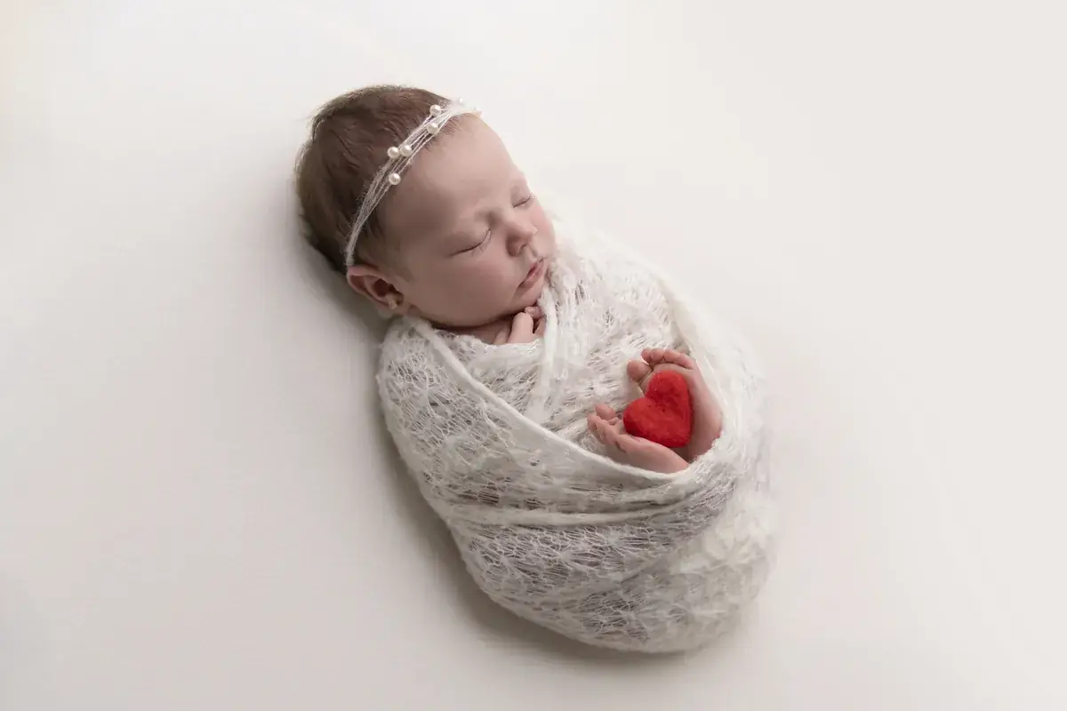 Newborn girl wrapped in cream lace knit, holding a tiny red felt heart — newborn photography by Gitty Reiss, Boro Park, Brooklyn